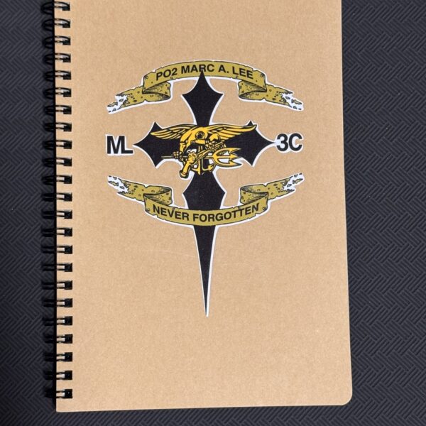 Beige spiral notebook with a large military emblem featuring a gold ribbon reading 'Never Forgotten' on a dark textured background.